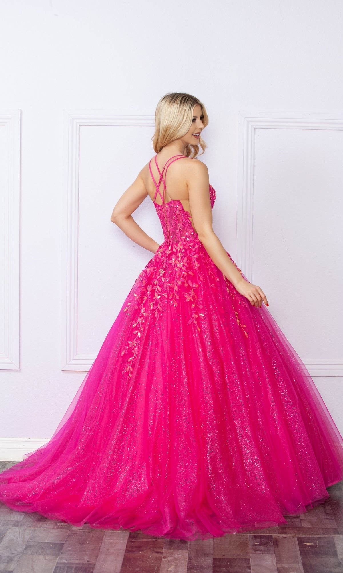 A woman stands with her back to the camera, wearing the Glitter-Tulle Long Prom Ball Gown H1271 in bright pink with floral accents and crisscross straps, its sparkling tulle skirt flowing to the floor against a white wall.